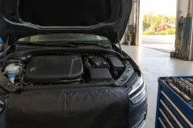 Öppen motorhuv över ett motorrum i en verkstadshall med verktygsvagn bredvid och porten öppen mot gården.