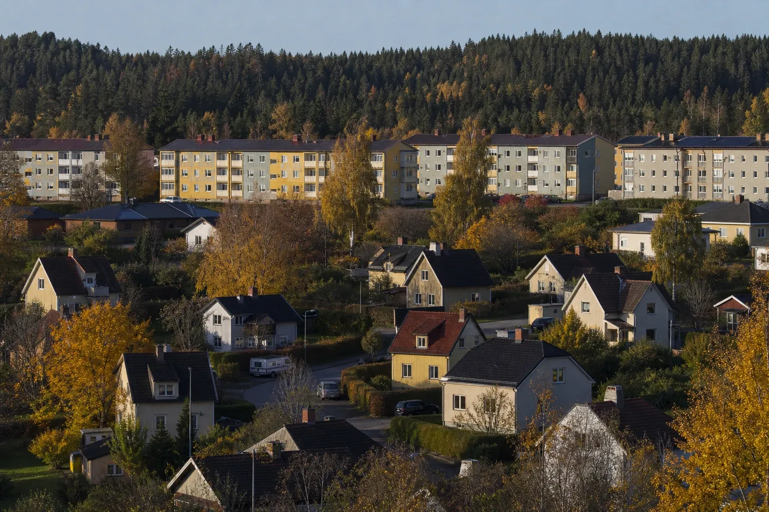 Utsikt över villakvarter och gula flerfamiljshus med skogsklädd ås bakom i höstfärger.