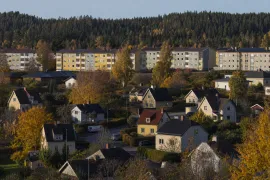 Utsikt över villakvarter och gula flerfamiljshus med skogsklädd ås bakom i höstfärger.