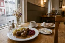 Tallrik med köttbullar, potatis och lingon på ett träbord vid ett fönster mot gatan.