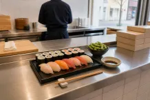 Fat med nigiri och maki på en stålbänk i ett sushikök, med kock i bakgrunden och staplade avhämtningslådor.