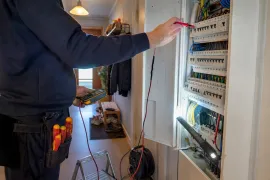 Elektriker mäter i en öppen elcentral i en hall, med skruvmejslar i bältet.